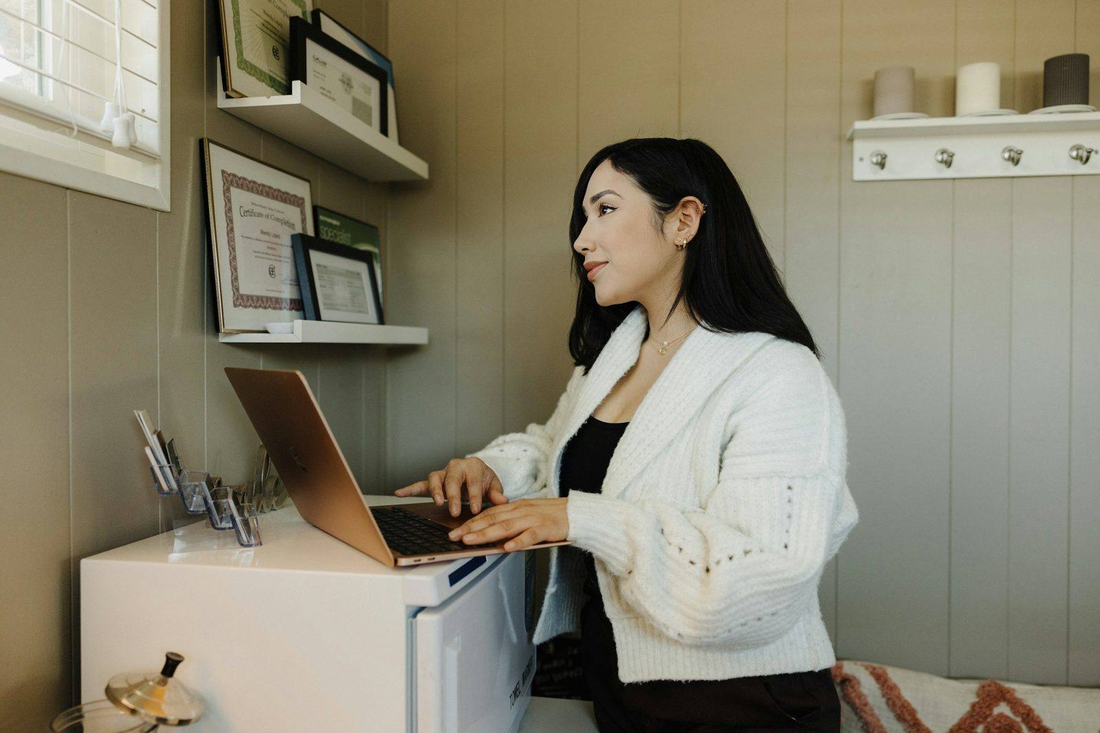 A woman typing on a laptop at a small home-office desk with framed certificates on the wall