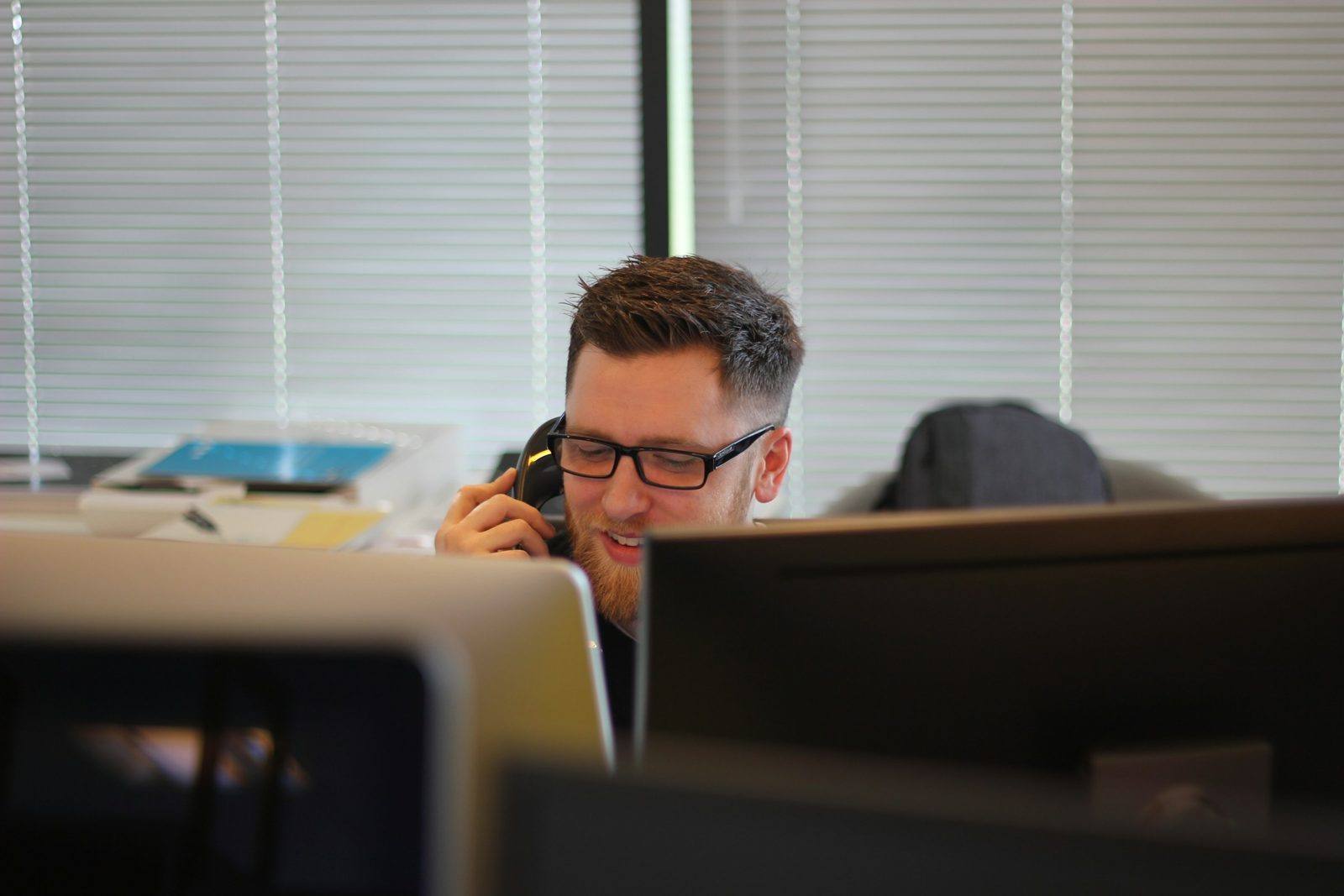 A man in glasses on the phone at his desk behind two computer monitors