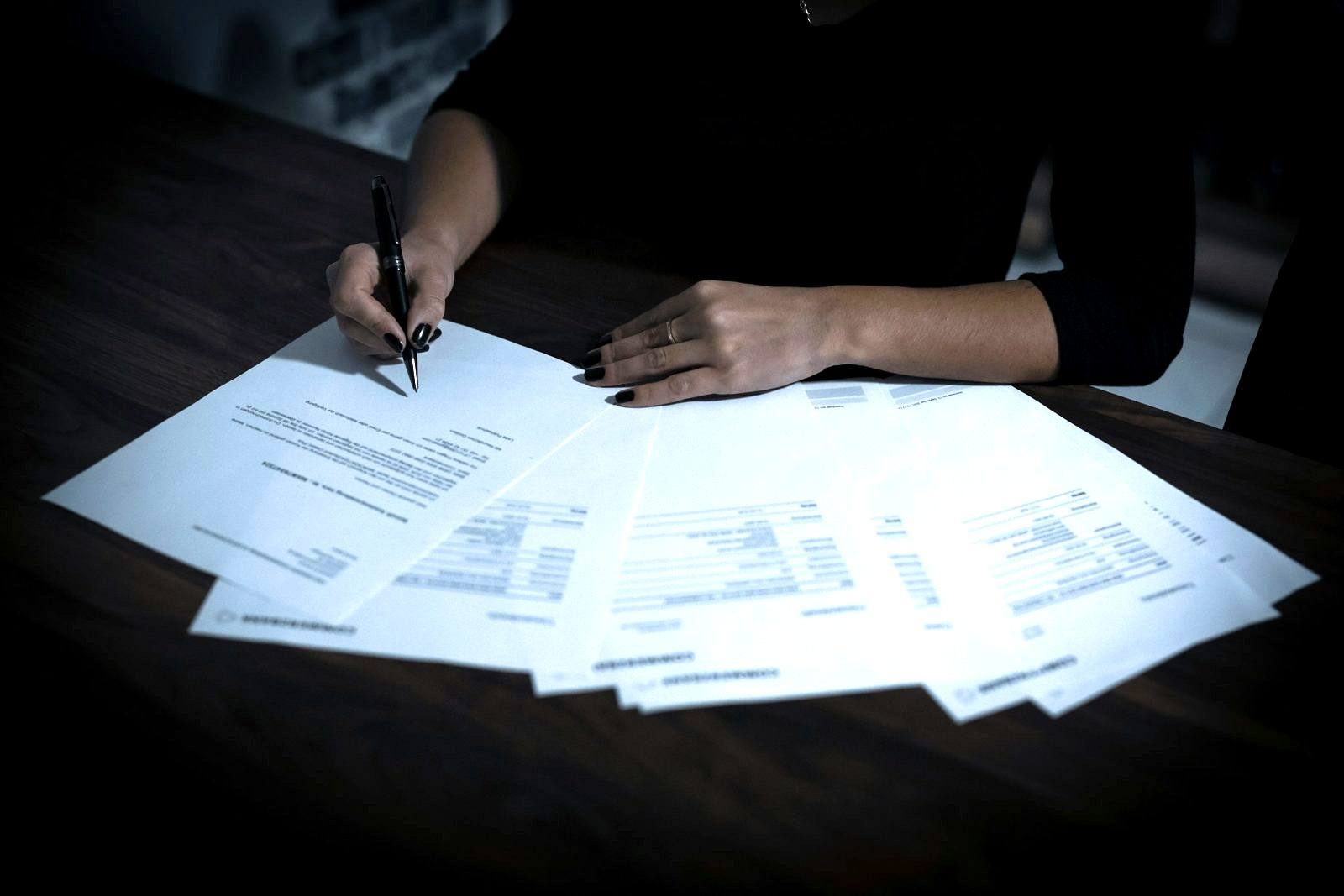 A hand signing a document, with several printed pages fanned out on a dark table